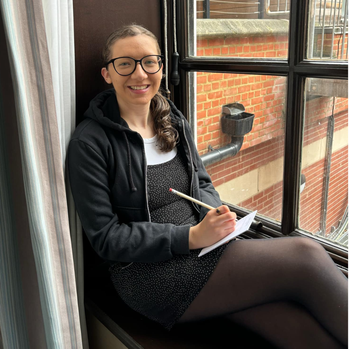 young woman on a windowsill writing with a pencil on a notepad